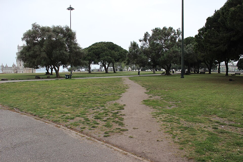 A dirt trail worn diagonally across a grassy park lawn, cutting the corner where two paved paths meet.